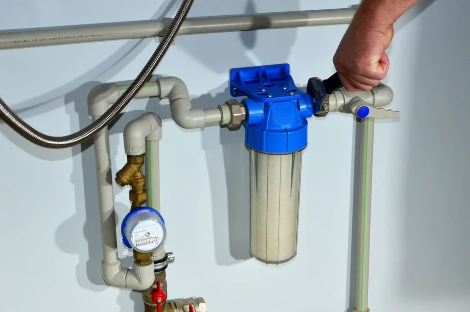 Technicien en tenue de travail professionnelle effectuant une maintenance routinière sur un adoucisseur d'eau résidentiel installé sous un évier ou dans une cave, en train de vérifier les paramètres ou d'effectuer un entretien annuel. Le technicien porte des gants de protection et manipule les composants de l'adoucisseur avec professionnalisme dans un environnement domestique. L'image doit montrer clairement le matériel caractéristique d'un adoucisseur (réservoir, raccordements) et transmettre la notion de service régulier et professionnel auprès d'un client.