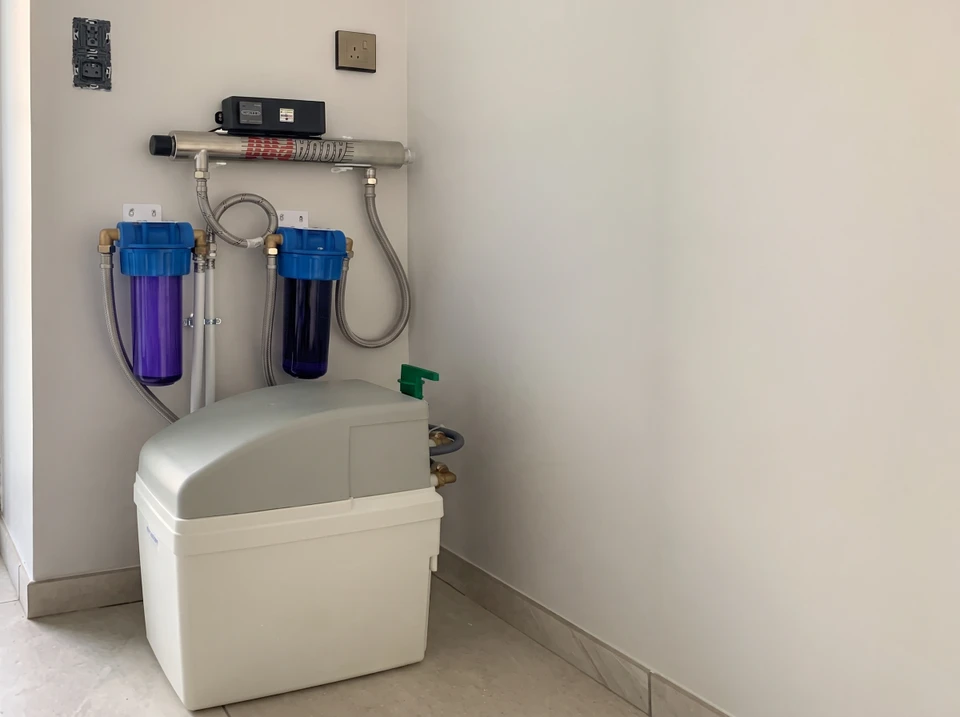 Technicien en tenue de travail professionnelle effectuant une maintenance routinière sur un adoucisseur d'eau résidentiel installé sous un évier ou dans une cave, en train de vérifier les paramètres ou d'effectuer un entretien annuel. Le technicien porte des gants de protection et manipule les composants de l'adoucisseur avec professionnalisme dans un environnement domestique. L'image doit montrer clairement le matériel caractéristique d'un adoucisseur (réservoir, raccordements) et transmettre la notion de service régulier et professionnel auprès d'un client.